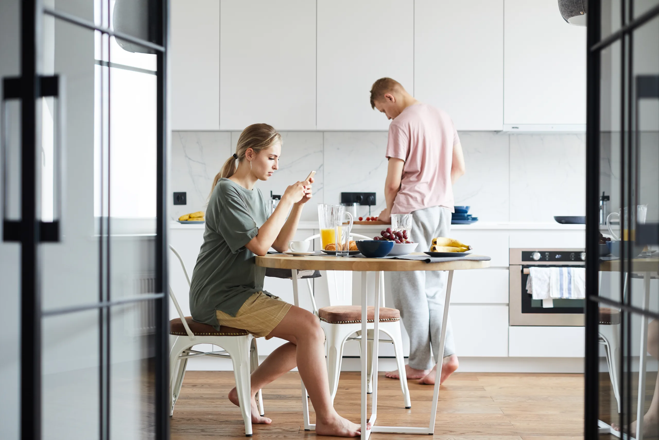 A woman sitting at a kitchen table looking at her phone while someone prepares food behind her